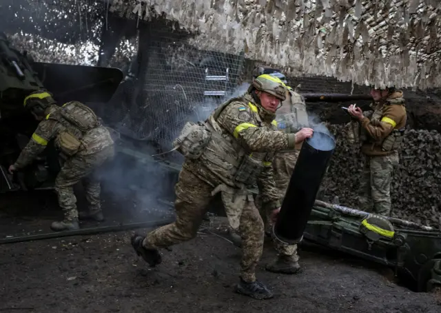 Украинский солдат в военной форме защитного цвета и бронежилете с желтыми наклейками несет двумя руками гильзу от артиллерийского снаряда. Из гильзы идет дым