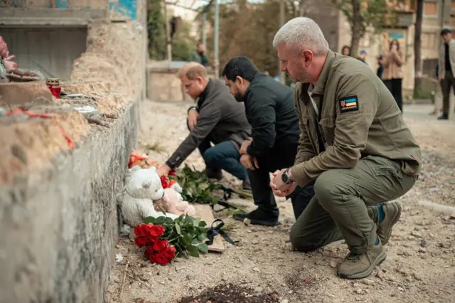 Принц Гарри, Тимур Ткаченко и человек в военной форме сидят на корточках у стены. На земле видны цветы и детский плюшевый медвеженок