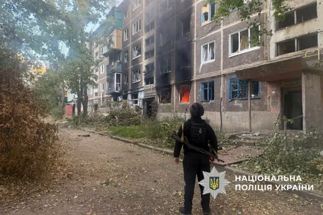 Военный в каске и бронежилете с автоматом стоит на фоне поврежденного жилого дома, в котором продолжается пожар