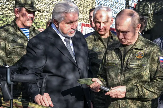 Путин в военной форме рядом с министром обороны РФ держит в руках пистолет. на груди у него герб России и надпись Путин В В