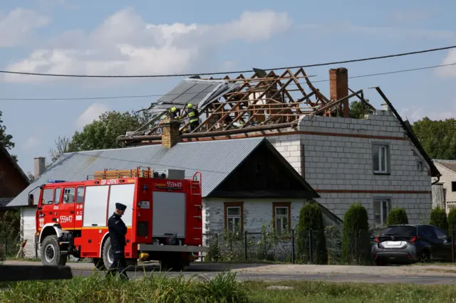 Пожарная машина и сотрудник силовых органов на фоне белого кирпичного дома с разрушенной крышей