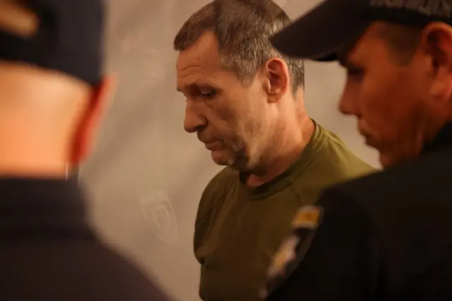 Подозреваемый в зале суда на фоне двух охранников. Он коротко стрижен, волосы с проседью. На нем зеленая футболка
