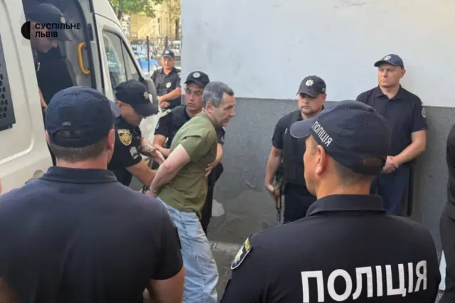 Подозреваемого выводят из машины в наручниках