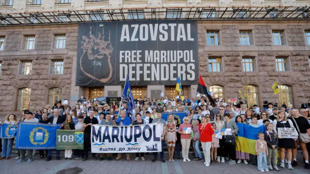 Митингующие позируют с плакатами и украинскими сине-желтыми и красно-черным флагами, на большом плакате над ними написано по-английски "Азовсталь - освободите защитников Мариуполя"