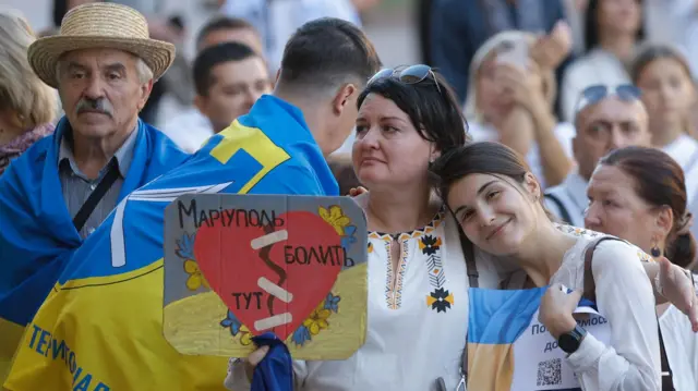 Пожилой мужчина в соломенной шляпе с украинским флагом на плечах, более молодой мужчина, завернувшийся во флаг, сфотографирован со спины, зрелая женщина в вышиванке и улыбающаяся девочка с украинским флагом и плакатом