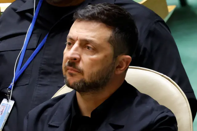 يرتدي زيلينسكي بدلة سوداء وقميصاً أسود دون ربطة عنق، تظهر الصورة الجزء العلوي من جسده أثناء جلوسه في مقر الأمم المتحدة.