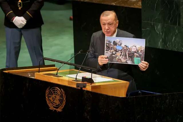إردوغان يرتدي بدلة رسمية رمادية اللون، ويقف متحدثاً على منصة للأمم المتحدة ويحمل بيديه صورة لحشد من الفلسطينيين في غزة يحملون أوانٍ فارغة وينشدون الطعام.