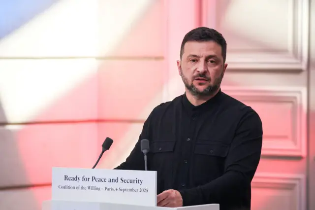 Владимир Зеленский во время пресс-конференции в Париже. Он стоит за трибуной в темной рубашке на фоне двери, подсвеченной розовым.