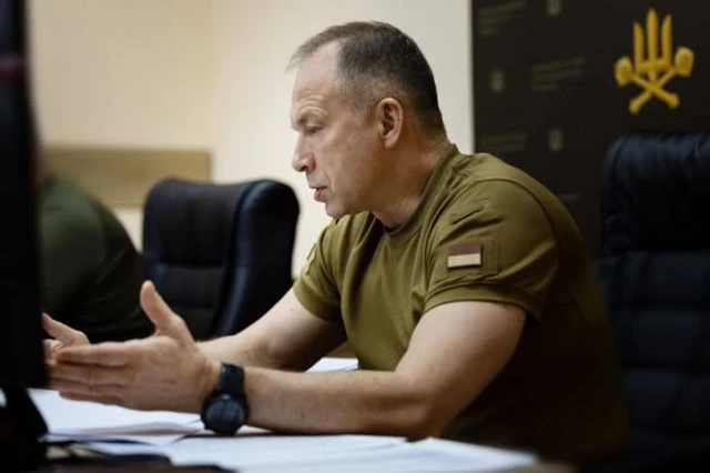 Сырский в военной футболке сидит за столом, он изображен в профиль, на руке у него видны часы