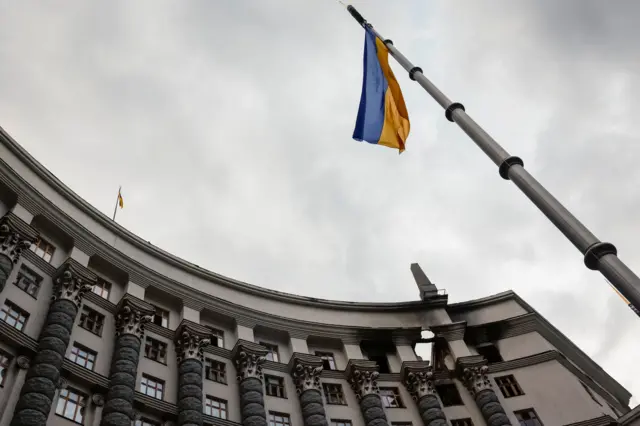 Здание Кабинета министров Украины, пострадавшее от обстрела. Вид снизу: флагшток с украинским флагом и здание Кабмина, в котором на верхних этажах видна пробоина