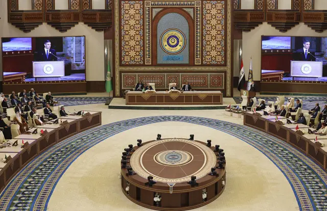 قاعة يجتمع فيها عدد من القادة العرب يغلب على ألوانها البني والبيج، وأمام كل وفد علم بلاده، وهناك شاشتان كبيرتان يظهر عليها المتحدث. وفي الوسط مقاعد للدولة المستضيفة.