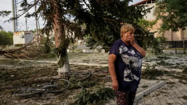 Женщина прикрывает рукой рот, под хвойным деревом лежат на земле два велосипеда
