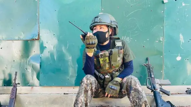 ပြောက်ကျားယူနီဖောင်းဝတ်ပြီး သံခမောက်ဆောင်း စကားပြောစက်ပြောနေတဲ့ အမျိုးသမီးတစ်ဦး ထိုင်နေတာကို မြင်ရ၊ သူ့ဘေးမှာ သေနတ်တလက်ထောင်ထား