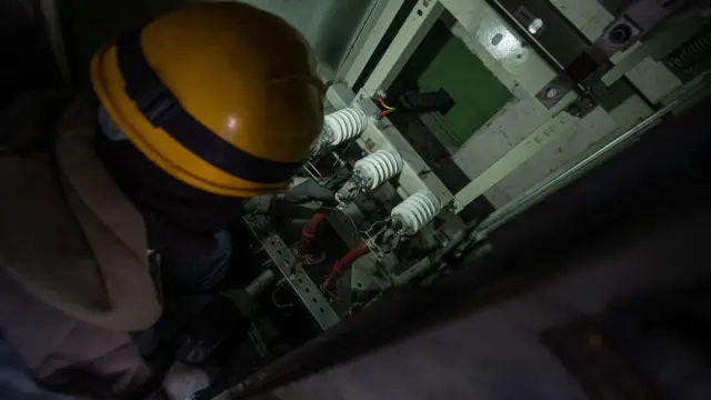 Рабочий в каске сфотографирован со спины, смотрит на фарфоровые изоляторы в темном помещении
