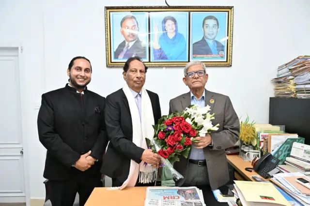 বিএনপি নেতা মির্জা ফখরুল ইসলাম আলমগীরের হাতে ফুলের তোড়া তুলে দিচ্ছেন অধ্যাপক আবু সাইয়িদ, পাশে আরো একজন দাঁড়িয়ে আছেন