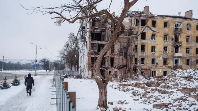 Руины желтой пятиэтажки под снегом, слева от фотографа удаляется человек, снятый со спины