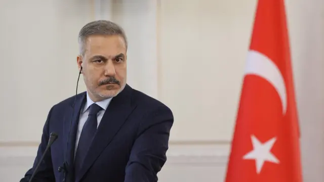 هاکان فیدان، وزیر خارجه ترکیه با کت و کراوات سورمه‌ای نشسته جلوی پرچم ترکیه مقابل میکروفن