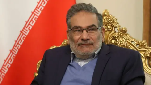 علی شمخانی با کت سیاه، پولیور آبی و پیراهن راه‌راه، نشسته روی مبل طلایی جلوی پرچم ایران