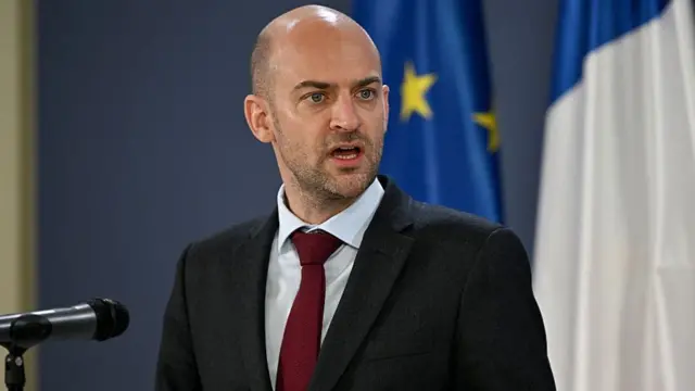 ژان نوئل بارو، وزیر خارجه فرانسه، با کت سیاه، پیرهن سفید و کراوات زرشکی ایستاده مقابل میکروفن