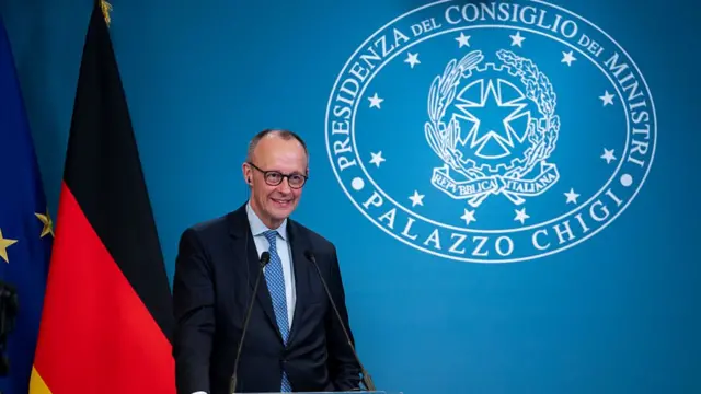 فردریش مرتس، صدراعظم آلمان، با کت سیاه و کراوات آبی ایستاده مقابل پرچم آلمان و پشت تریبون