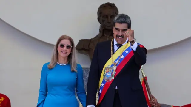 Imagen de Nicolás Maduro y Cilia Flores. Maduro levante el puño, viste de traje y tiene puesta la banda presidencial. Cilia Flores lleva un traje azul y gafas de sol.