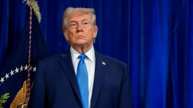 Trump em pé, com olhar sério em frente a bandeira e fundo azul