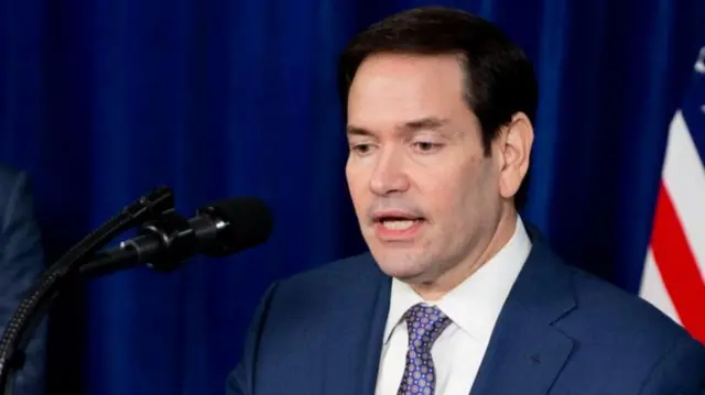 Rubio falando ao microfone, com bandeira dos EUA atrás