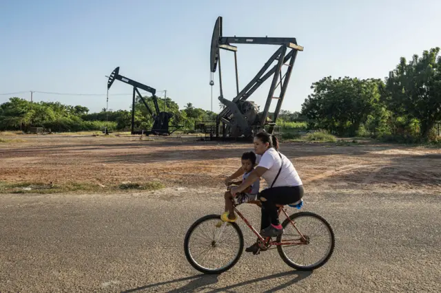 Mulher leva criança em bicicleta duante de guindastes em campo de extração de petróleo em Cabimas, na Venezuela, em 18 de dezembro de 2025