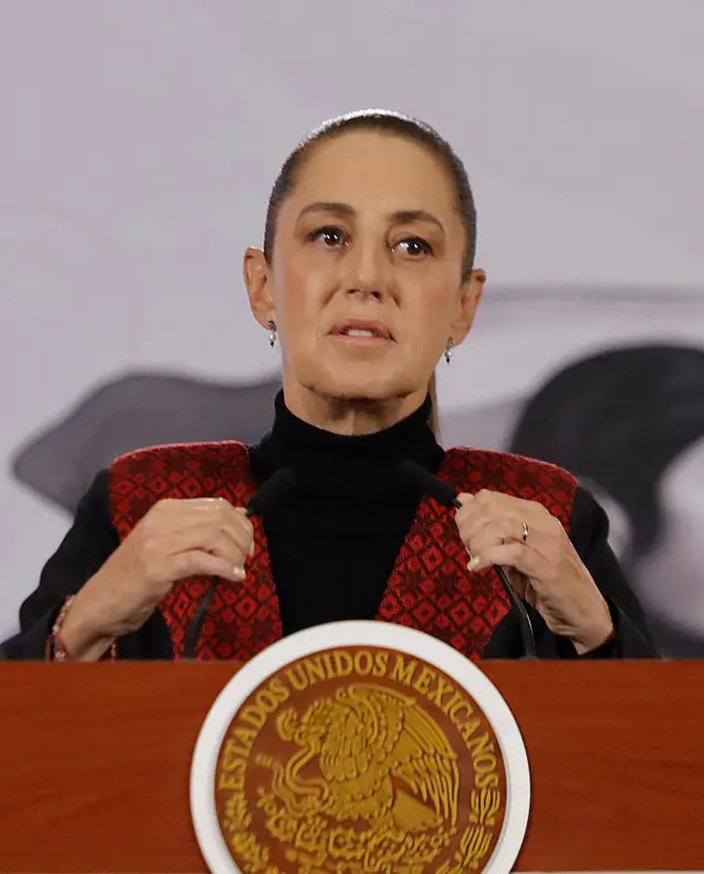 رئيسة المكسيك، كلوديا شينباوم تصفف شعرها للخلف بإحكام ليتدلى كـذيل الحصان.، وترتدي سترة سوداء.