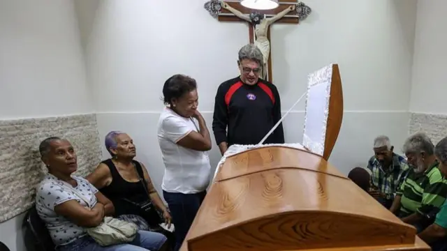 Un hombre y una mujer miran un ataúd rodeado de personas que acompañan el velatorio.