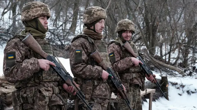 Трое солдат в камуфляже с нашивками в виде украинского флага стоят, направив дула автоматов вниз