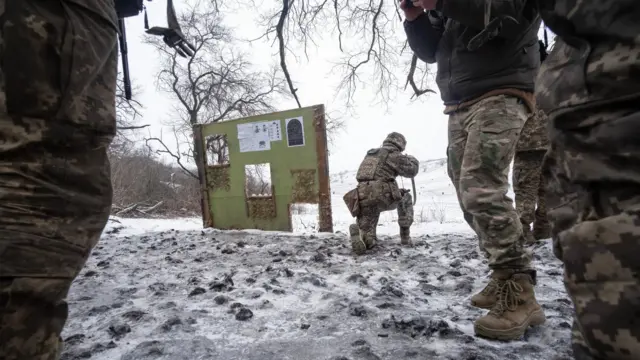 Сфотографированный со спины человек в камуфляже целится из винтовки, стоя на одном колене, на переднем плане несколько человек, одетых в камуфляжную форму, стоят, их лица за кадром
