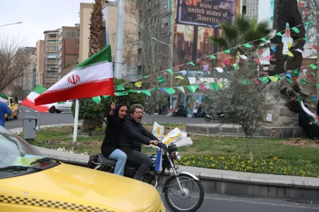 মোটরসাইকেলে করে যাচ্ছেন একজন পুরুষ ও একজন নারী যার হাতে ইরানের একটি পতাকা