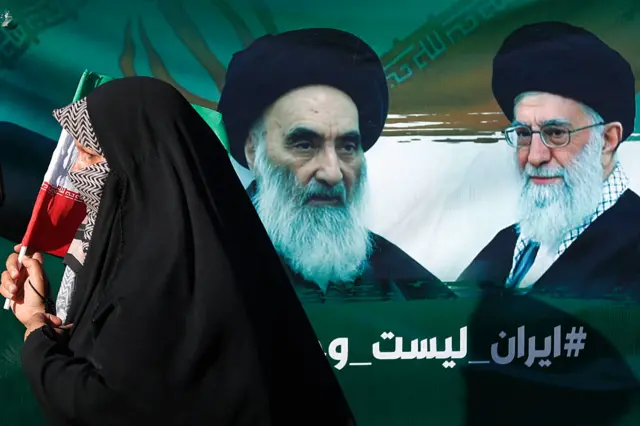 امرأة عراقية تمر أمام صورة للمرجع الشيعي الأعلى في العراق، آية الله علي السيستاني، والمرشد الأعلى للجمهورية الإسلامية الإيرانية، آية الله علي خامنئي، خلال مسيرة في حي الكاظمية ببغداد في 21 يونيو/حزيران 2025، احتجاجًا على الضربات الإسرائيلية على إيران.