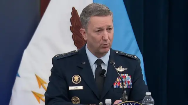 General Caine resmi üniformasıyla kürsüde konuşuyor.