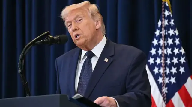 Prezida Donald Trump ariko arashikiriza ijambo yambaye ikoti, inyuma yiwe hari idarapo rya Amerika.