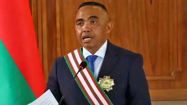 Koloneli (Col) Michael Randrianirina arimo gusoma ijambo (imbwirwaruhame) yambaye ikositimu iriho umwitero wo ku rutugu, n’umudari ku ikoti. Iruhande rwe hari ibendera rya Madagascar.