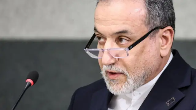 Arakçi mikrofona konuşuyor, yakın gözlüklerinin üzerinden ileri bakıyor.