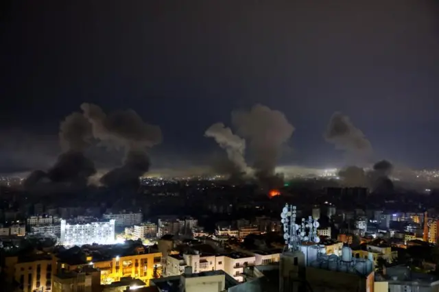 صورة ليلية لمدينة بيروت مضاءة بأضواء المباني. وفي الأفق، تتصاعد ست سحب دخانية ضخمة من المدينة، ويبدو أن إحداها تحترق.