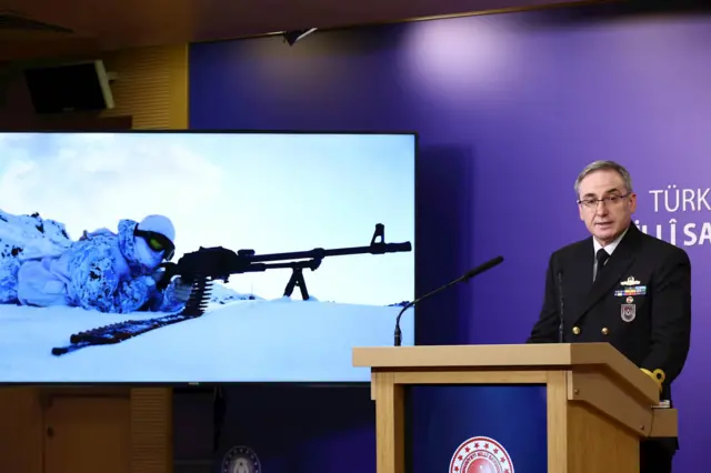 Zeki Aktürk üniformasıyla kürsüde konuşuyor, yanında bir perdeye beyaz kamuflajlı bereli karlar üstüne uzanmış bir asker sniper'a bakıyor.