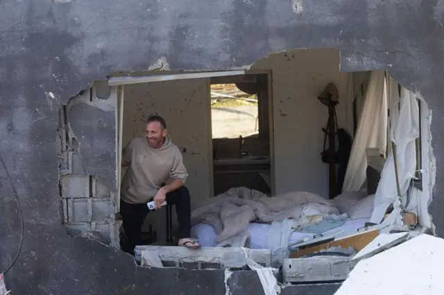 হামলায় ক্ষতিগ্রস্ত একটি বাড়ির ভাঙ্গা দেয়াল ঘেঁষে হাসিমুখে দাঁড়িয়ে আছেন একজন ব্যক্তি।