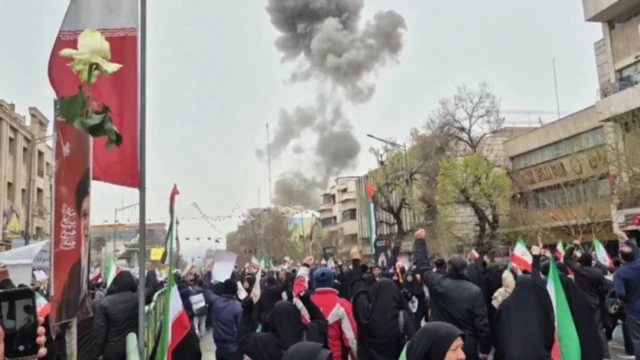 Толпа людей с иранскими флагами на фоне дыма