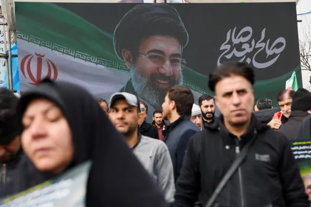 يسير الناس بجوار لافتة تحمل صورة المرشد الأعلى الإيراني الجديد، مجتبى خامنئي، خلال مراسم جنازة قادة الجيش الإيراني الذين قُتلوا في غارات جوية، في طهران، إيران، 11 مارس/آذار 2026