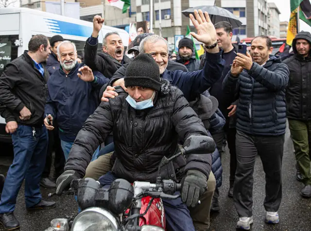 İran Cumhurbaşkanı Mesud Pezeşkiyan, bir motorsikletin arkasında el sallıyor, çevredekiler onun bu ilginç halinin fotoğrafını çekiyor.