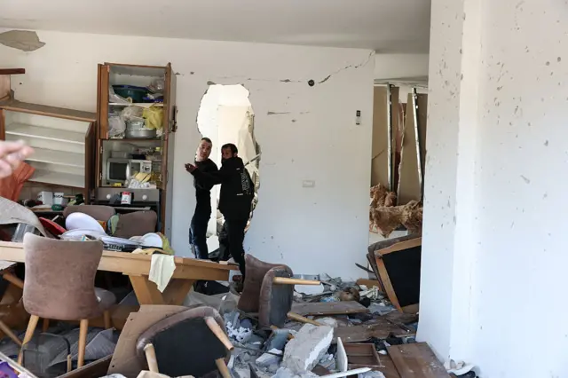 رجلان يتفقدان منزلاً متضرراً في موقع سقوط صاروخ في بلدة زارزير، بشمال إسرائيل، وأمامها طاولة وكراسي محطمة.