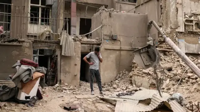 Ifoto iriko umuntu ari kuri telefone , inyuma yiwe hari inzu yasambuwe n'igisasu