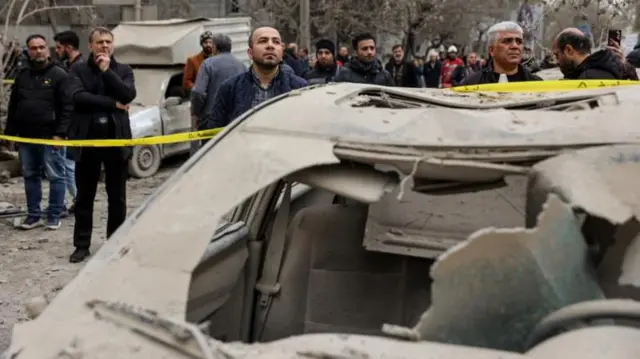 হামলায় বিধ্বস্ত একটি গাড়িকে ঘিরে মানুষ ক্ষয়ক্ষতি দেখছেন