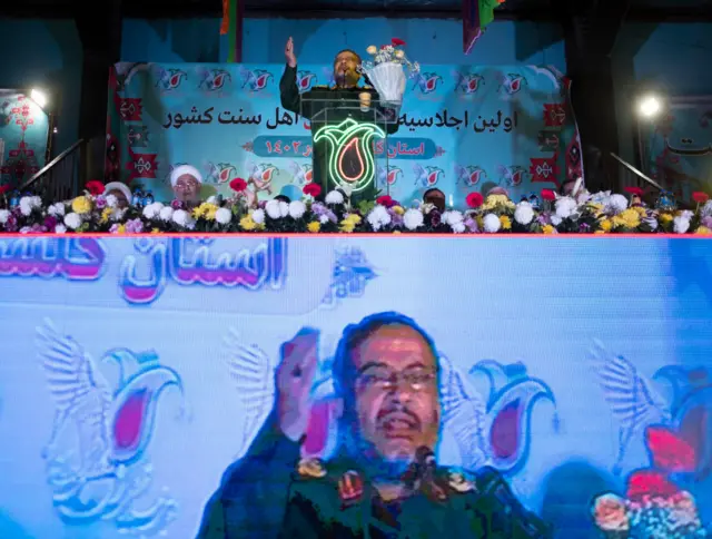 süleymani çiçeklerle dolu bir sahnede konuşma yapıyor, altta da büyük ekrandan konuşması gösteriliyor.