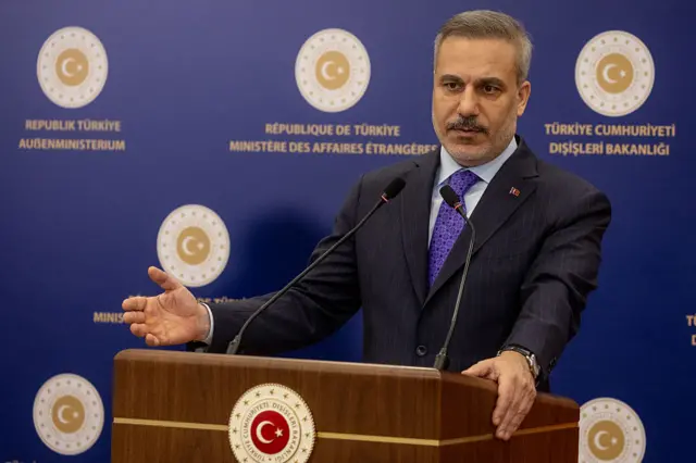 Hakan Fidan bir kürsünün arkasında bir elini yukarı kaldırmış konuşuyor. Arkada üzerinde Dışişleri Bakanlığı'nın logosu bulunan bir platform var.