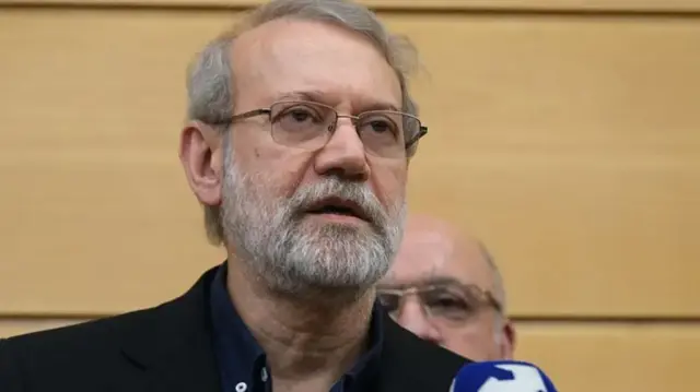 Laricani gözlüklü ve sakallı önündeki mikrofona konuşuyor. Arkasında sarı ahşap bir panelin önünde gözlüklü bir başka erkeğin yüzü görünüyor.